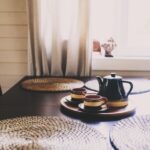 A close-up of a dark, rustic dining table near a window. The table has placemats and a tea set in the center.