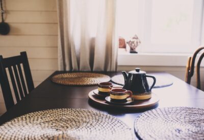 A close-up of a dark, rustic dining table near a window. The table has placemats and a tea set in the center.