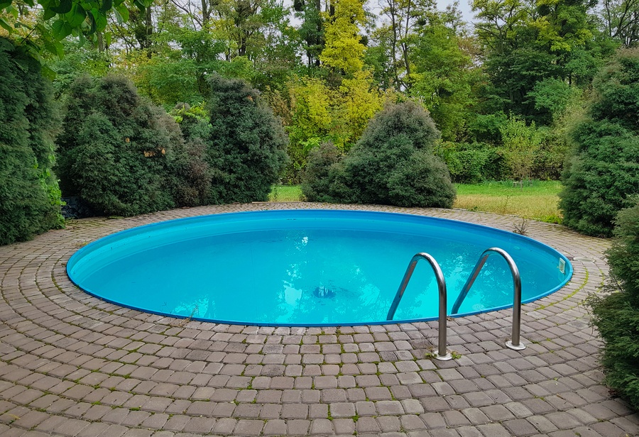 A small circular pool surrounded by brown brick pavers and lush green trees and bushes on a cloudy day.