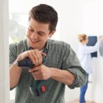 A young man smiling while using a power drill on a doorframe. A young woman tests paint colors on a wall behind him.