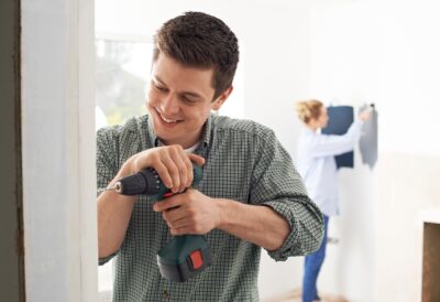A young man smiling while using a power drill on a doorframe. A young woman tests paint colors on a wall behind him.
