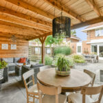 Wooden chairs surround a round table opposite a gray wicker couch in a outdoor room with wood-paneled walls and ceiling.