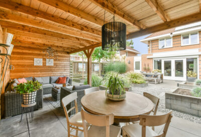 Wooden chairs surround a round table opposite a gray wicker couch in a outdoor room with wood-paneled walls and ceiling.