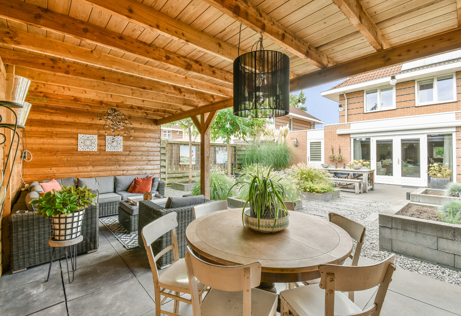 Wooden chairs surround a round table opposite a gray wicker couch in a outdoor room with wood-paneled walls and ceiling.