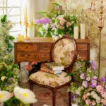 A vintage floral chair surrounded by lush flowers, books, and candles in a bright, elegant room.