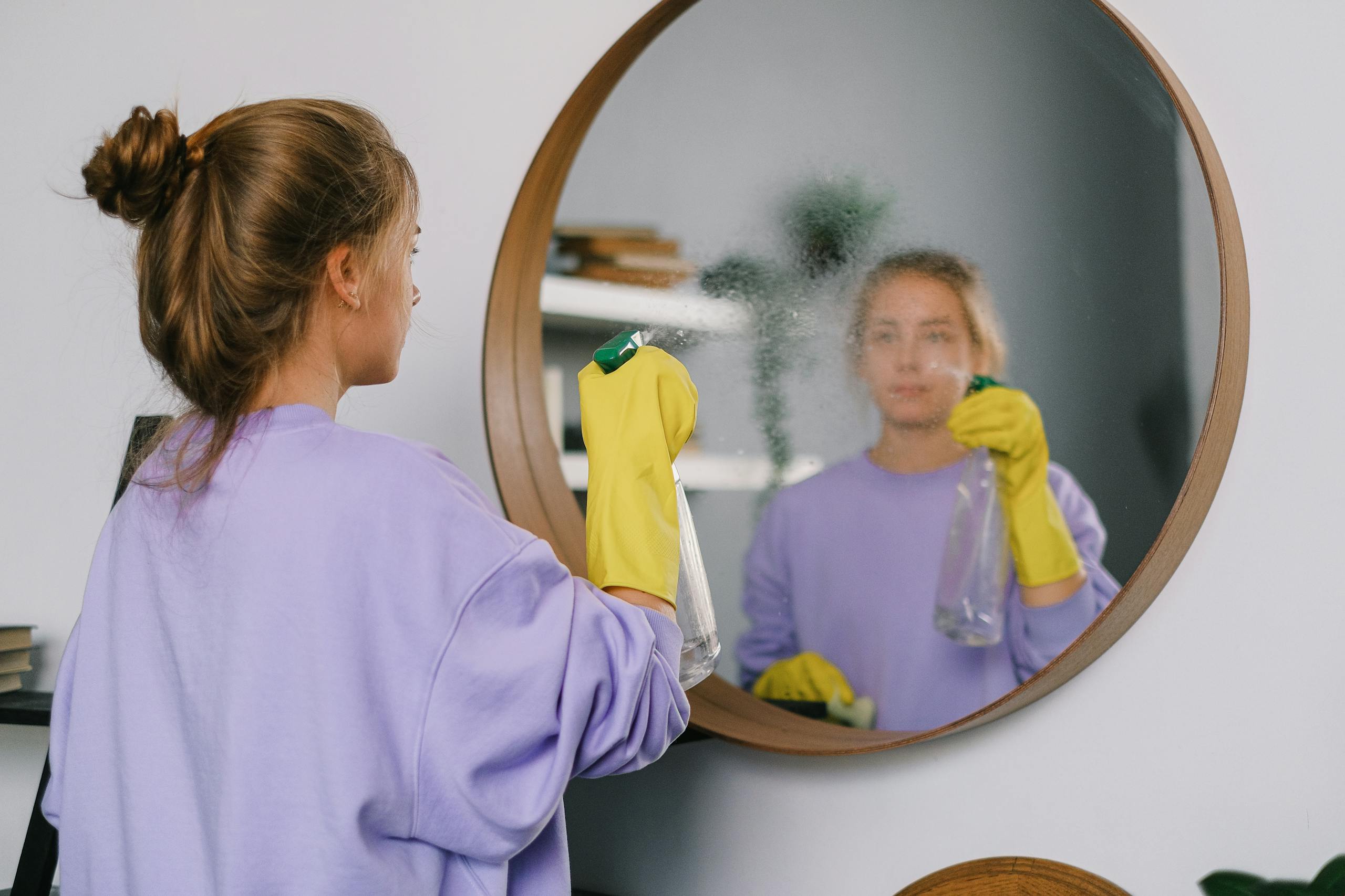 Woman in yellow gloves cleaning a mirror indoors using a spray bottle.